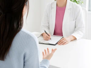 Doctor in a white coat signs a clipboard while speaking with a patient across a white table.