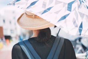 Person seen from behind wearing a straw sun hat and blue backpack under a geometric-patterned umbrella on a sunny urban street.