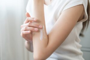 Person applying lotion to their forearm, focusing on skincare routine in a bright room, wearing a white top.