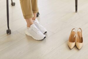 Person wearing white knit sneakers and rolled-up khaki pants stands near a pair of beige slingback heels on a light wooden floor.