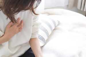 Person sitting on a bed, hand pressed to their chest as if in discomfort or pain in a bright, cozy room.