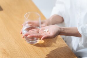 Person holding two white pills on an open palm with a glass of water on a wooden table nearby.